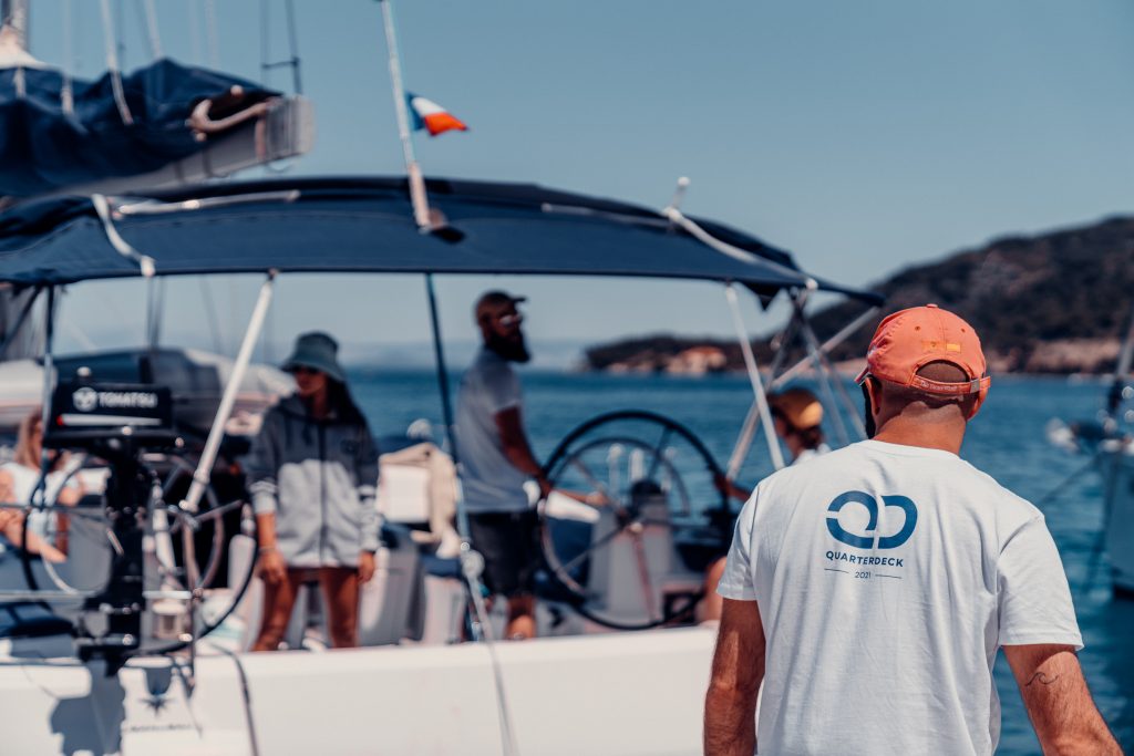 A skipper and crew members on the helm deck of a sailboat navigating open waters on a sunny day.