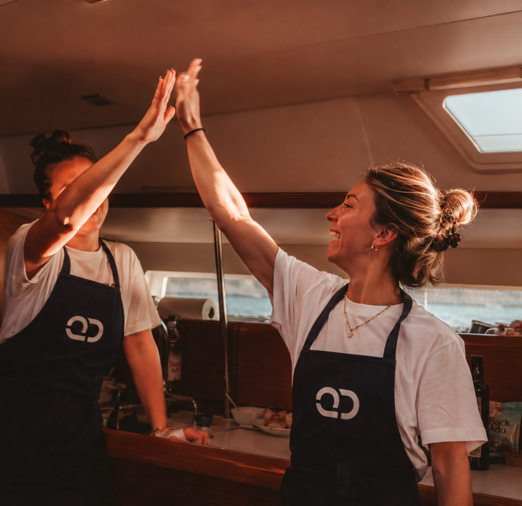 Two smiling female yacht chefs wearing navy blue Quarterdeck branded aprons giving each other a high-five inside a sun-lit wooden yacht galley.