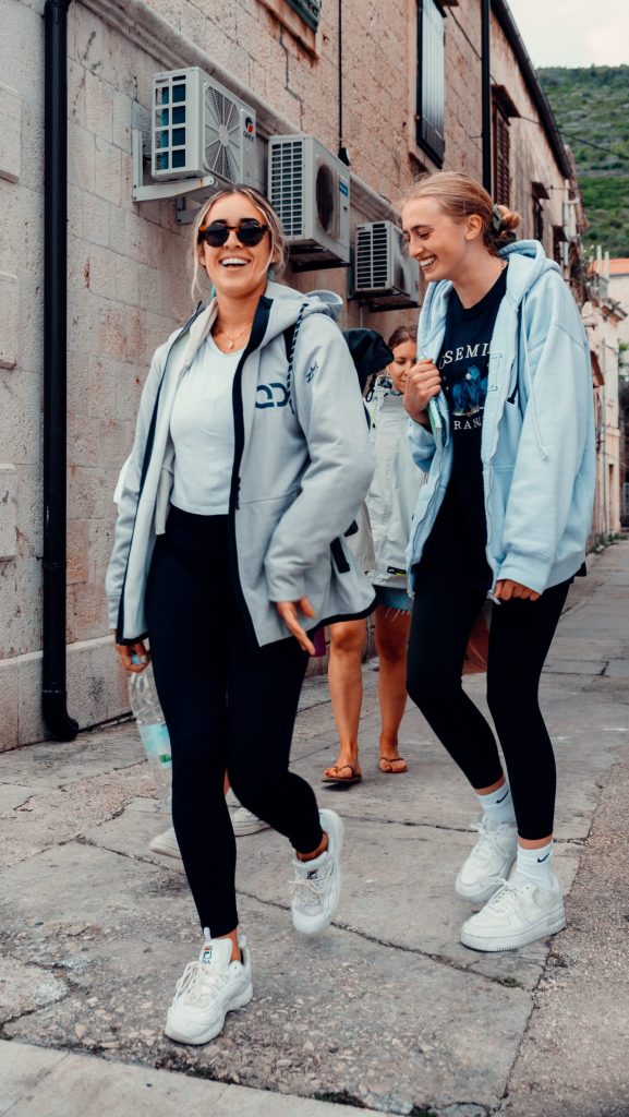 Two smiling female yacht host students from the Quarterdeck Host Academy walking outdoors on a cobblestone Mediterranean street, one wearing a branded Quarterdeck jacket.