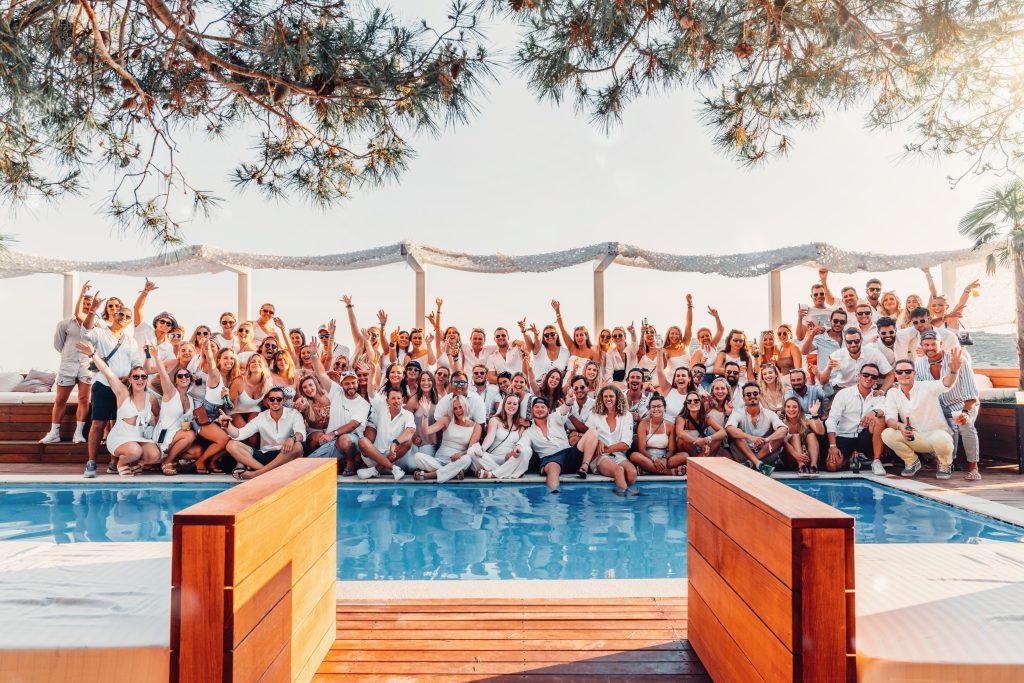 A large, energetic group of Quarterdeck flotilla crew members dressed in white, cheering together behind a bright blue swimming pool at a Mediterranean beach club.