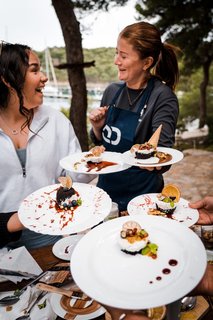 A smiling female chef/host in a Quarterdeck-branded dark blue apron holds two elaborate plated desserts, while a guest holds another. A variety of unique, artistically presented dishes are shown in the foreground and background, with blurred trees and boats. The logo is visible.