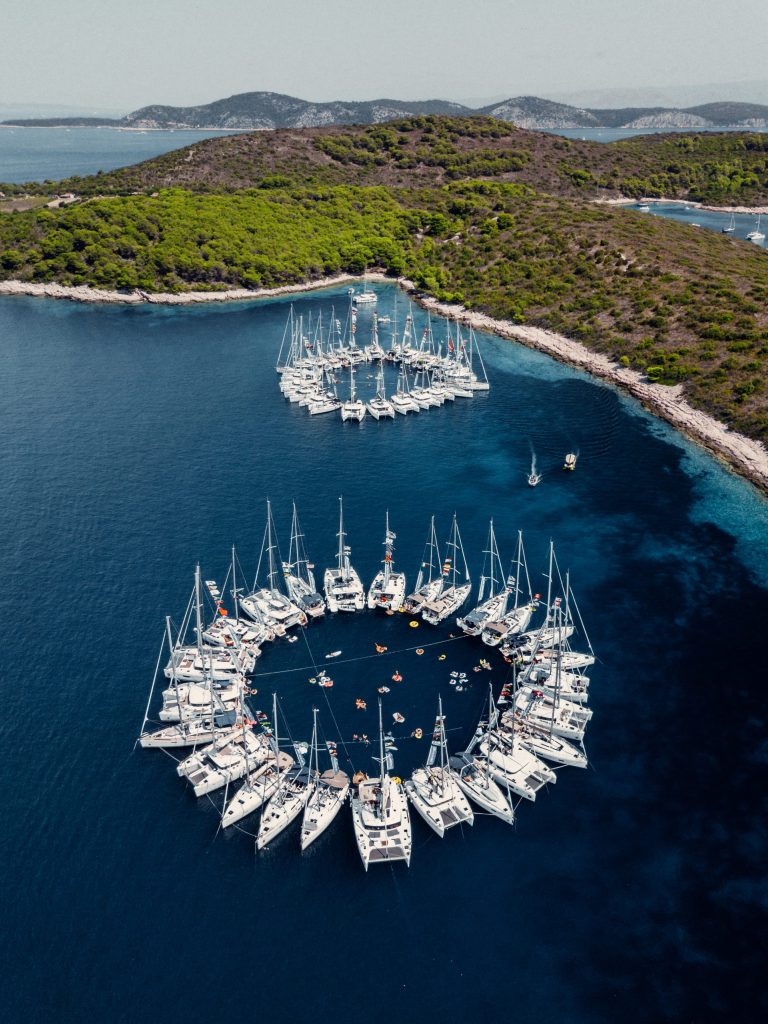 High-angle aerial drone photograph showing two massive circles formed by dozens of yachts (catamarans) and other boats, linked together in the deep blue sea near a lush green island in Croatia. Small specks (people swimming) are visible in the center of both large circles. One circle is closer to the foreground.