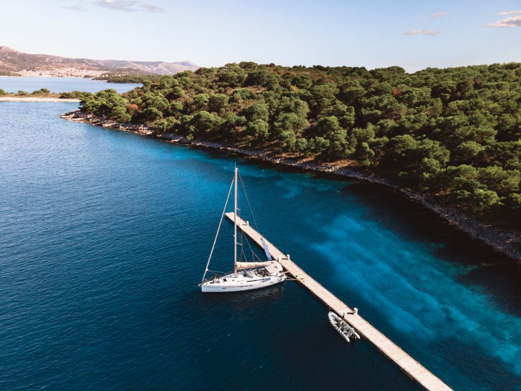 Aerial view of a sailboat docked at a long wooden pier along a lush green coastline with clear turquoise water.