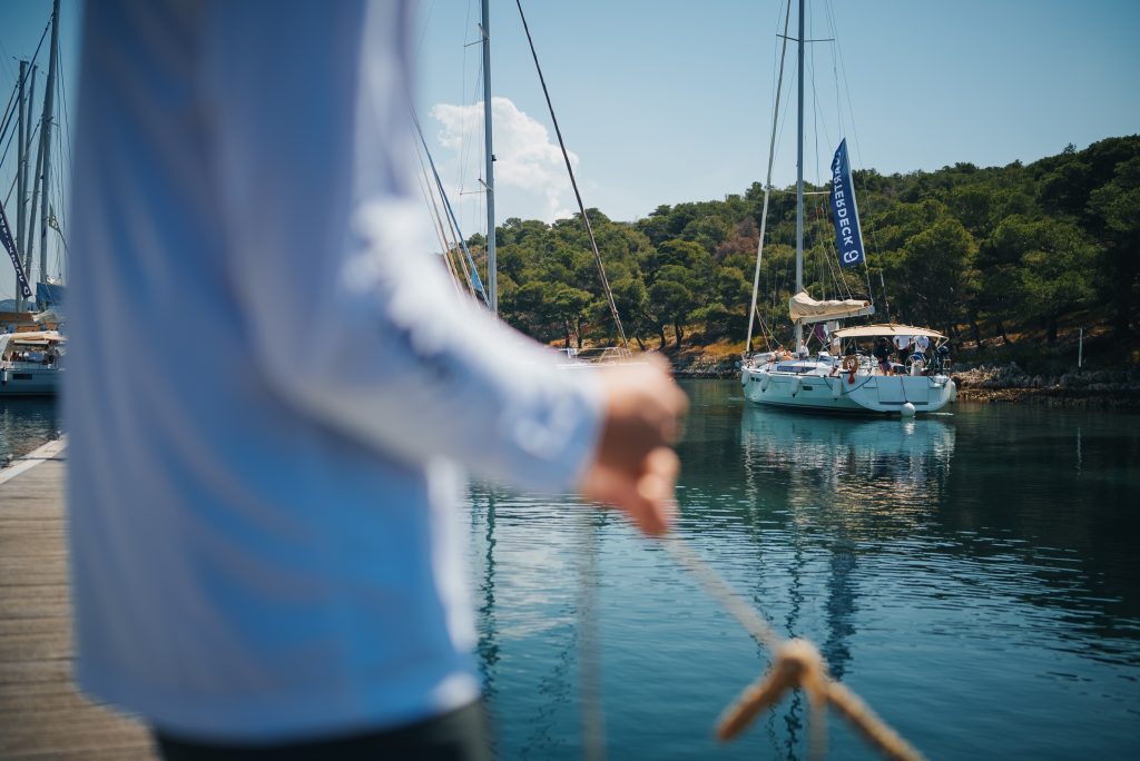 A blurred foreground of a yacht crew member holding a mooring line on a dock, looking out toward sailboats anchored near a tree-lined shore.