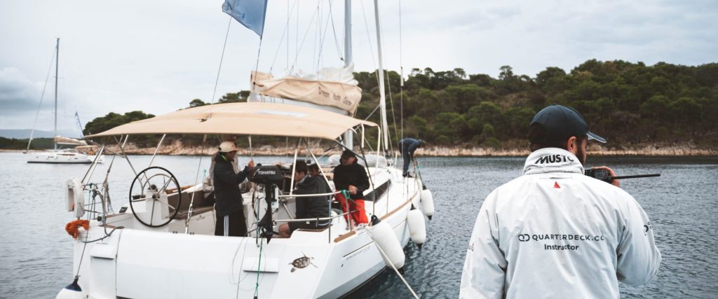 Quarterdeck instructor watching students maneuver a sailboat with a academy flag during a practical Skipper Academy training module.
